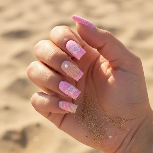 Hand with pink Aloha Clawture false press on nails and floral designs against a sandy background