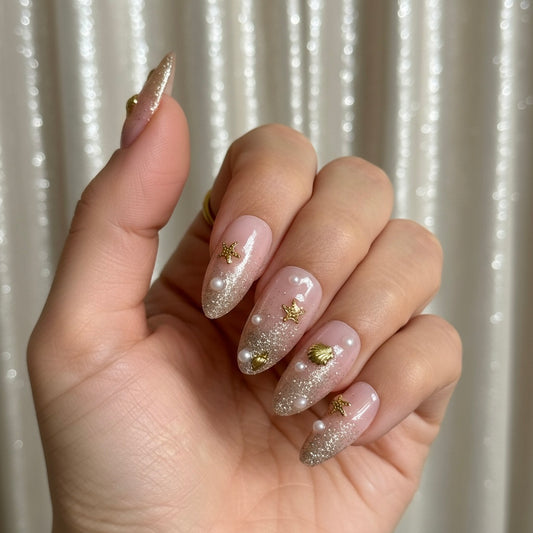 A close-up of a hand showcasing a manicure with Clawture Golden Shore Press on false nails long, stiletto-shaped nails decorated in a pink-to-clear glitter gradient, adorned with small white pearls and gold star accents.