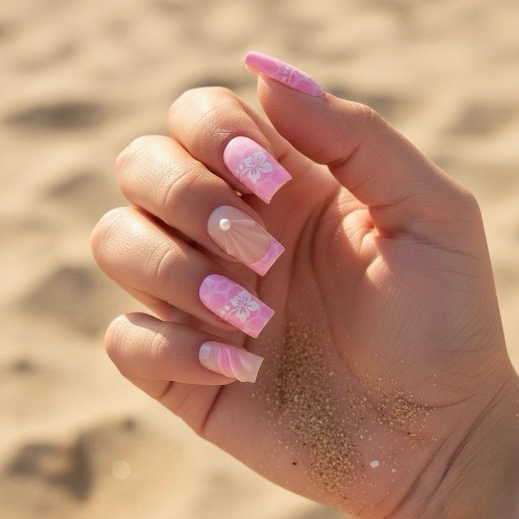 Hand with pink Aloha Clawture false press on nails and floral designs against a sandy background