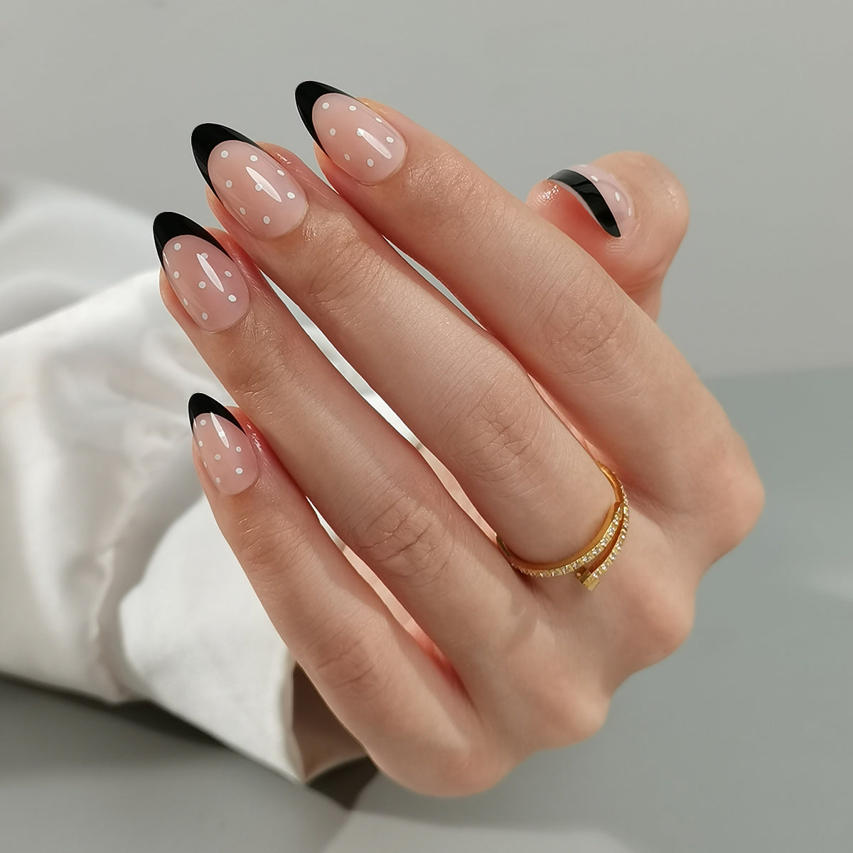 Black & Dotty Clawture press on nails: hand with almond nude nails, black French tips and a gold ring on a light grey background.