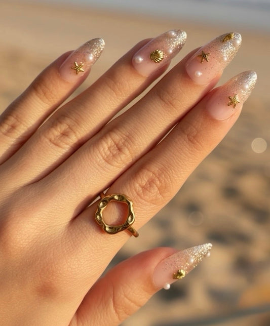 A close-up of a hand Clawture Golden Shore Press on false nails with long, glittery nude nails decorated with small gold star and shell motifs, wearing a gold ring with a textured open circle design. The background is a softly blurred sandy beach.