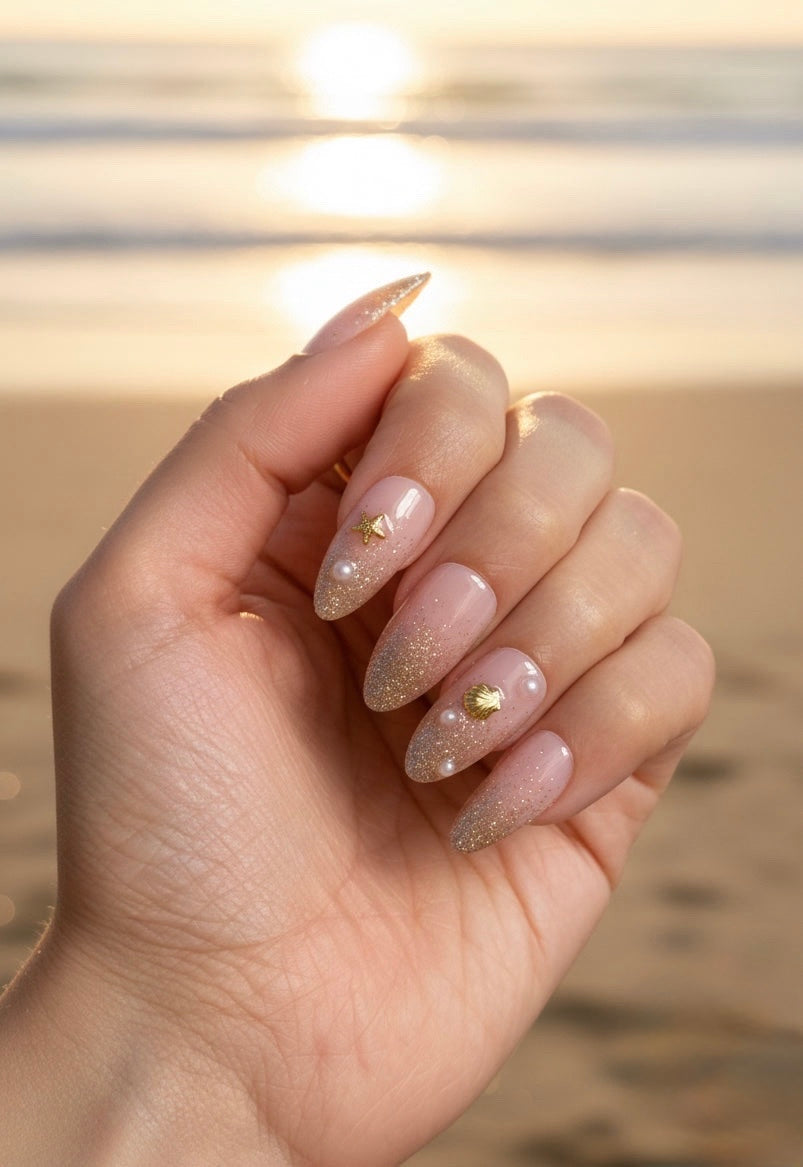 A close-up of a hand with Clawture Golden Shore Press on false nails long almond-shaped nails featuring a glossy nude pink base, fine gold glitter, and small gold heart and triangle nail art, posed against a blurred beach sunset background.