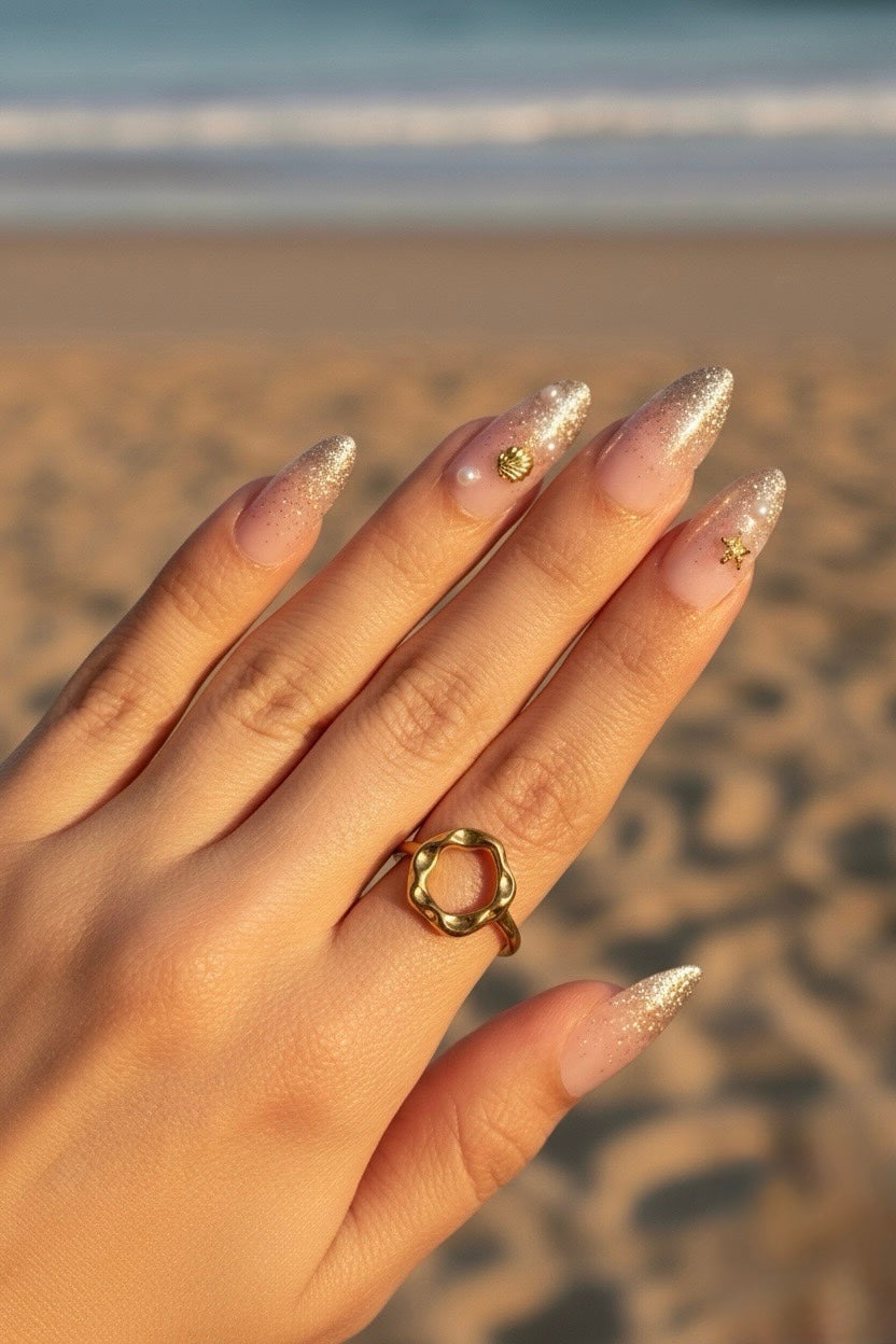 A close-up of a hand with Clawture Golden Shore Press on false nails glittery nude nails and small gold nail art, wearing a gold ring with a textured circular band, set against a blurred beach background.