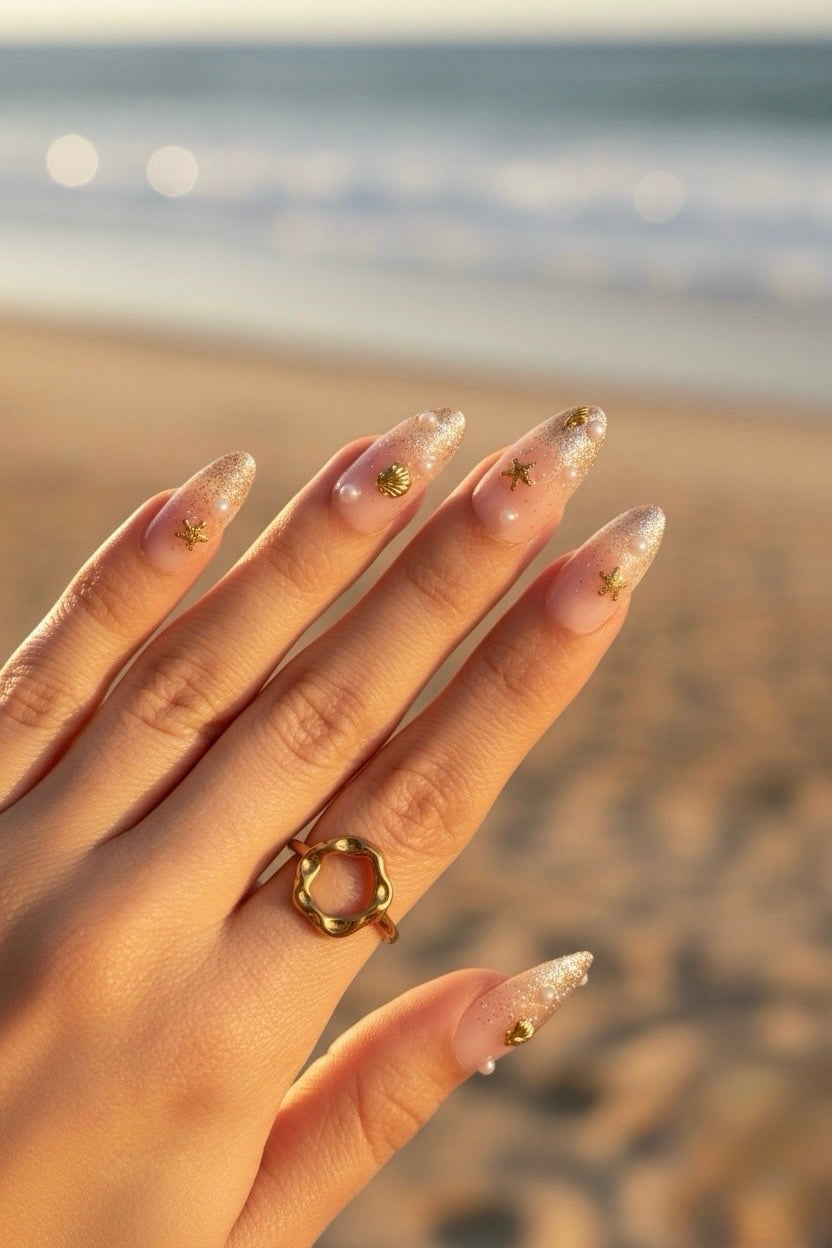 A close-up of a hand with Clawture Golden Shore Press on false nails long, glossy nude-pink nails decorated with small gold star motifs, wearing a gold ring, set against a blurred beach background.