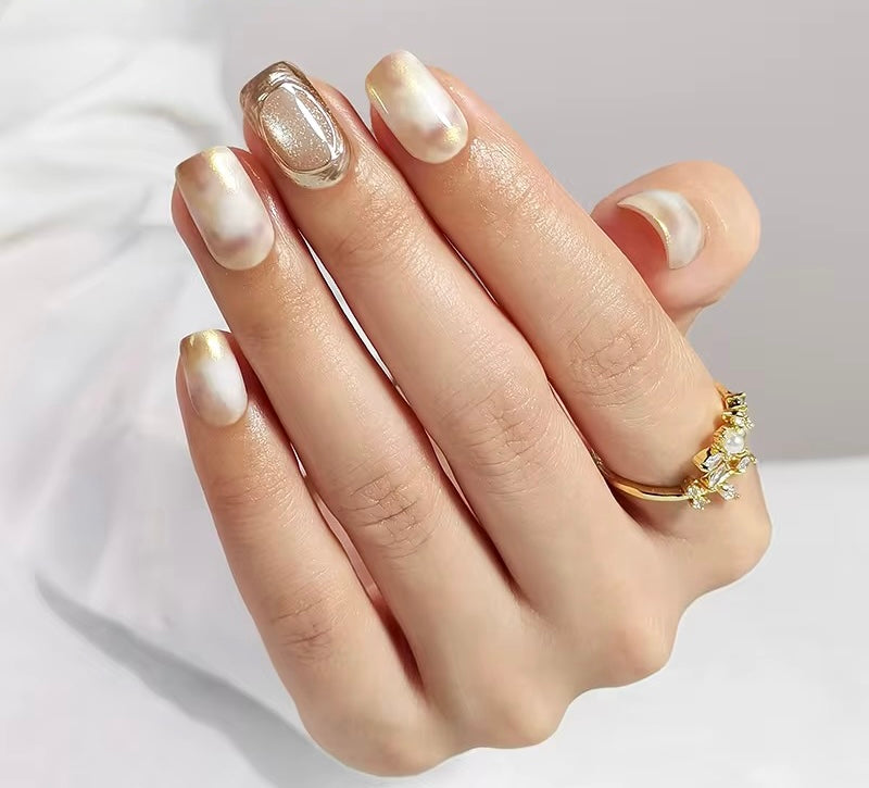 Hand with gold and white press on nails wearing a gold ring on a light background clawture