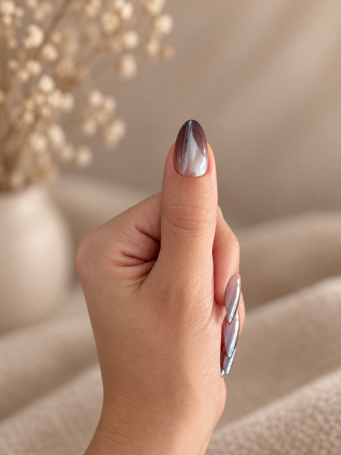 Cocoa Ombré Clawture press on nails: close-up of a hand with long almond-shaped brown chrome cat-eye nails set against a soft beige background with blurred dried flowers.