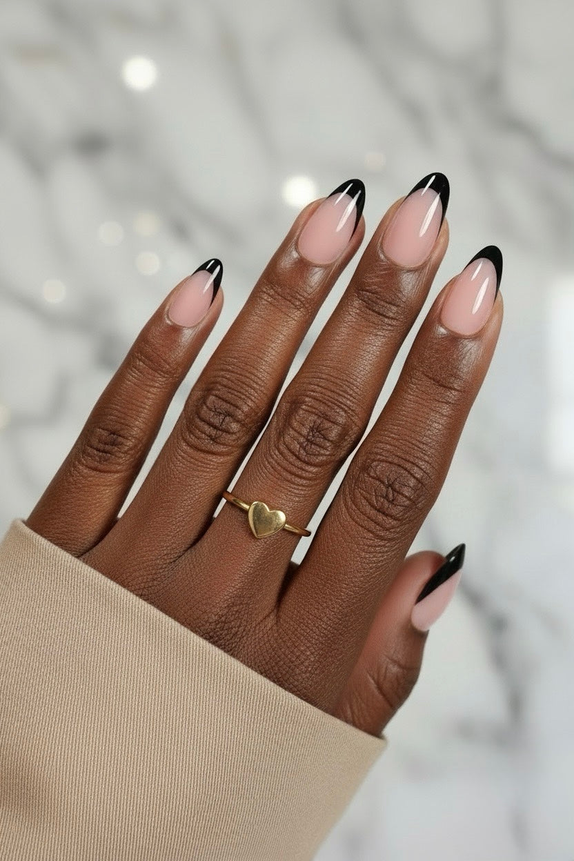 Hand with black swan clawture press on false nails beige nails and black tips, wearing a gold heart-shaped ring, against a blurred background.
